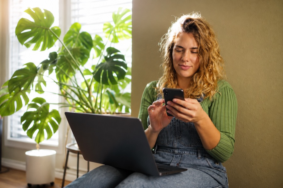 Mulher sentada perto da janela olhando o celular com o laptop no colo